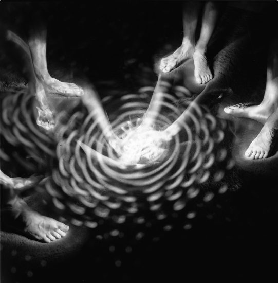 This black and white photograph shows a circle of bare feet, glowing white. They surround a strange spiral of white light.