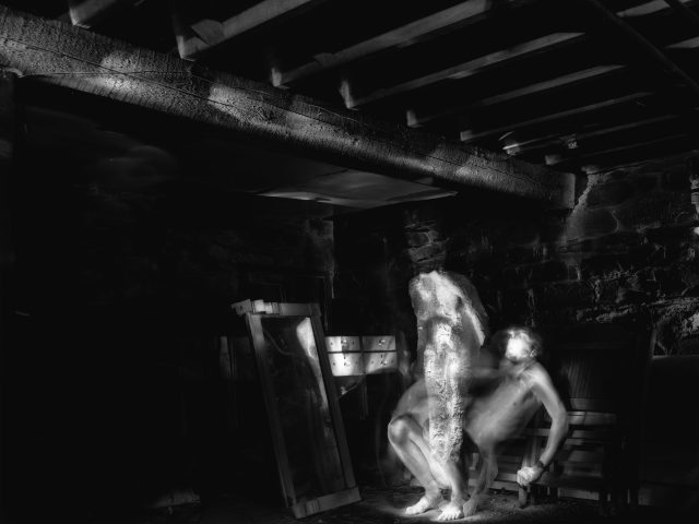 This black and white photograph shows the ghostly, naked figures of two men in a very dark room.