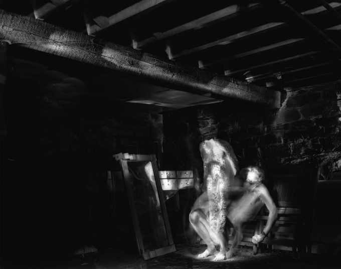 This black and white photograph shows the ghostly, naked figures of two men in a very dark room.