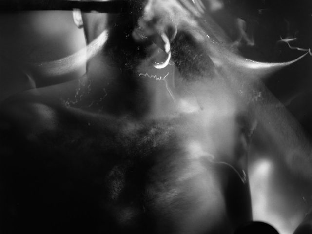 This black and white photograph shows the face of a man, close up. He is slightly blurred in motion and pale white light surrounds his head.