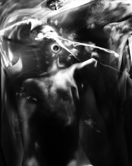 This black and white photograph shows the blurred figure of a naked person with a tube running out of their mouth. Bright flashes of light surround their head.