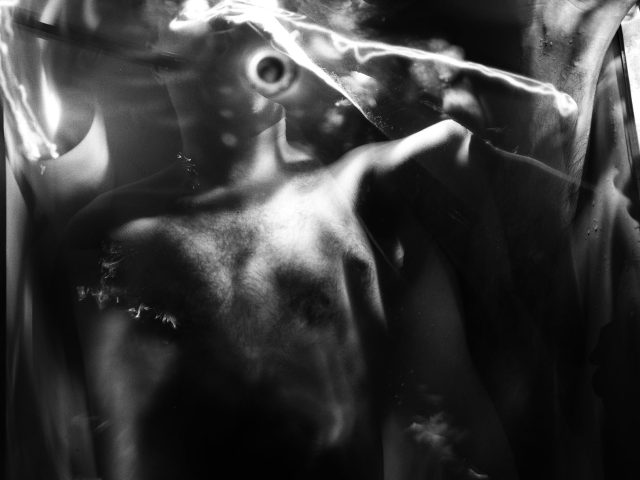 This black and white photograph shows the blurred figure of a naked person with a tube running out of their mouth. Bright flashes of light surround their head.