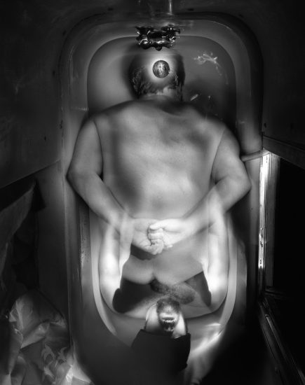 This black and white photograph shows the blurred figure of a man crouching in a bathtub, his figure half submerged. The ghostly silhouette of another figure is layered over his lower half.