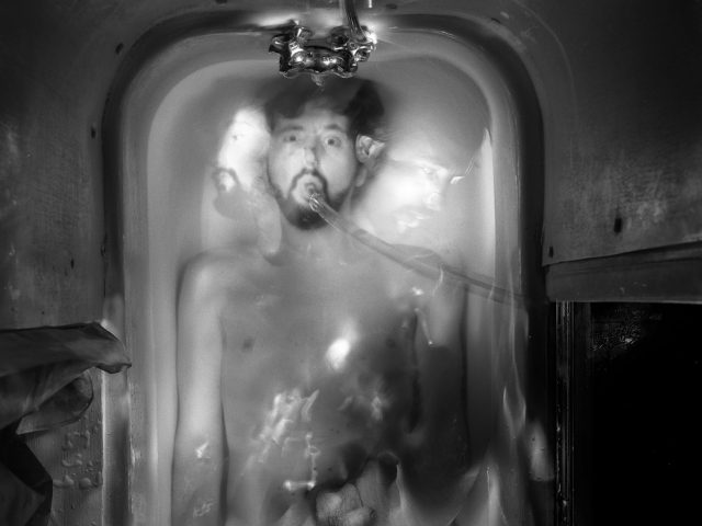 This black and white photograph shows a naked man lying half submerged in a bathtub, seen from above. The man has a tube running out of his mouth, outside the frame.