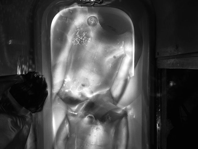 This black and white photograph shows glowing bodies lying cramped in a bathtub. They are seen from above, and the room around them is dark.