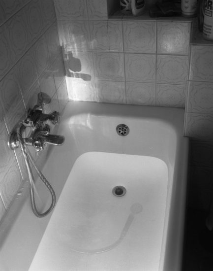This black and white photograph shows a bathtub full of water, dimly lit from above. A shadow is cast by the faucet onto the tile wall..