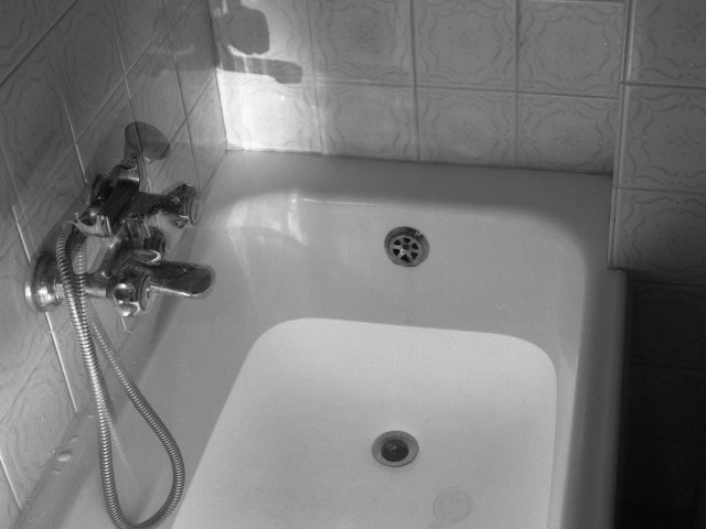 This black and white photograph shows a bathtub full of water, dimly lit from above. A shadow is cast by the faucet onto the tile wall..