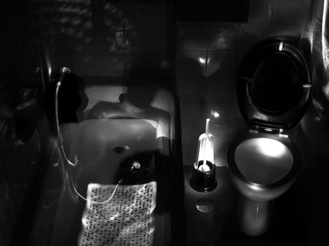 This photograph shows a dark bathroom. The tub is filled with water and it glows softly. The toilet lid beside it is open.