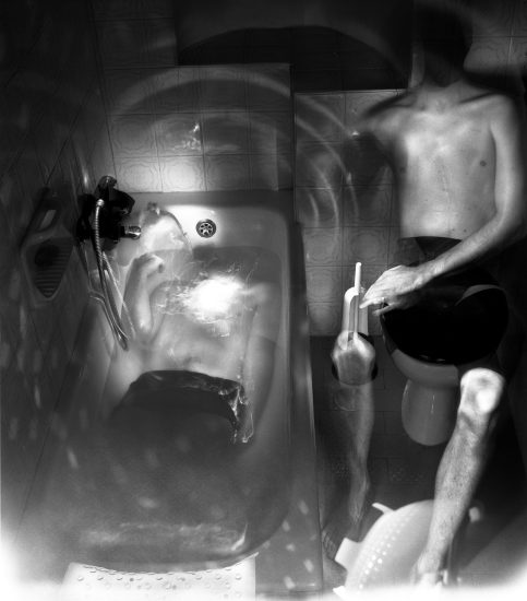 This photograph shows a bathtub, seen from above. The naked bodies of two men are seen, one lying in the tub and the other sitting next to him.