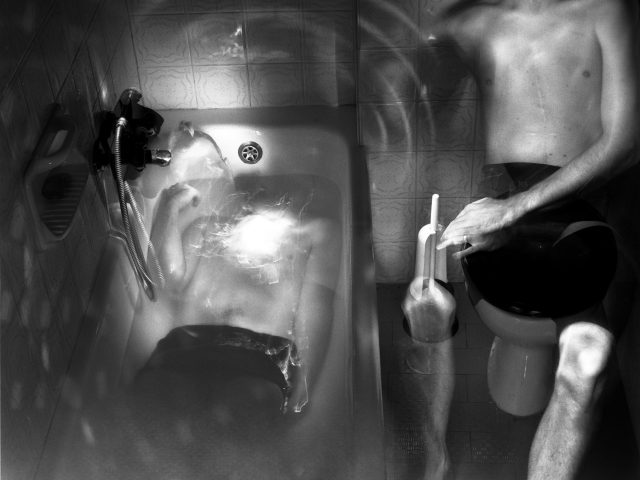 This photograph shows a bathtub, seen from above. The naked bodies of two men are seen, one lying in the tub and the other sitting next to him.