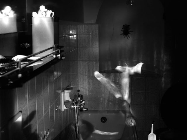 This photograph shows a dark bathroom space where the ghostly figure of a naked man sits on the toilet. His limbs are slightly blurred with motion.