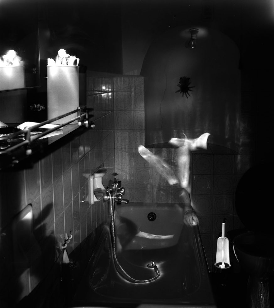 This photograph shows a dark bathroom space where the ghostly figure of a naked man sits on the toilet. His limbs are slightly blurred with motion.