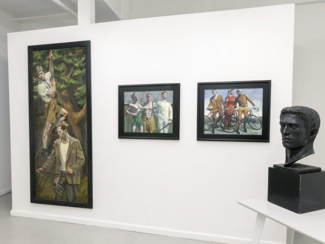 This photograph shows the walls of the gallery space where three framed paintings hang. On a table in the foreground, stands a black statue of a man's head from the neck up.