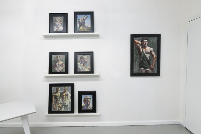 This photograph shows the walls of a gallery space where framed paintings of young men are hung or placed on thin shelves.