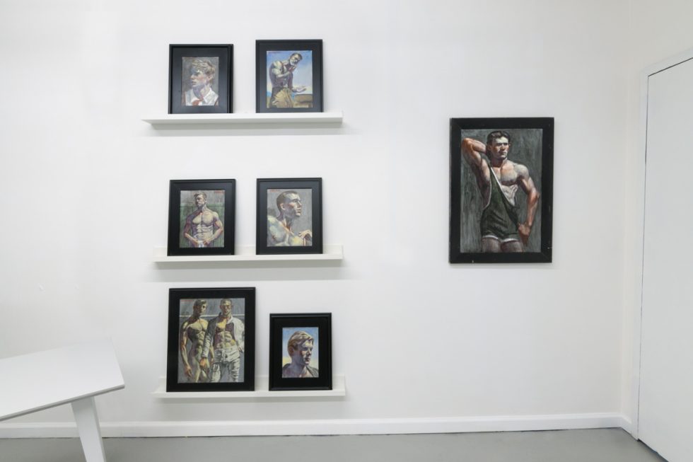 This photograph shows the walls of a gallery space where framed paintings of young men are hung or placed on thin shelves.