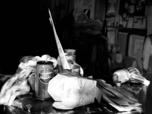 This photograph shows the surface of a table laden with assorted items including a Pepsi can, paint brushes, and Chinese food containers.