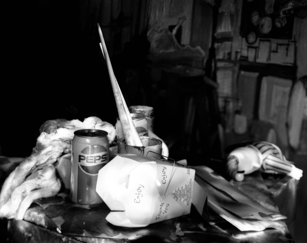This photograph shows the surface of a table laden with assorted items including a Pepsi can, paint brushes, and Chinese food containers.