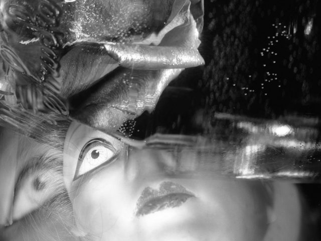 This black and white photograph shows the close up face of a doll. It's eyes are wide and white, looking up at the half hidden petals of a rose.