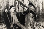 In this black and white image, a person wearing a hat poses in a forest by holding a vine hanging from a tree in each hand.