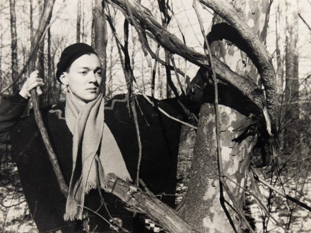 In this black and white image, a person wearing a hat poses in a forest by holding a vine hanging from a tree in each hand.