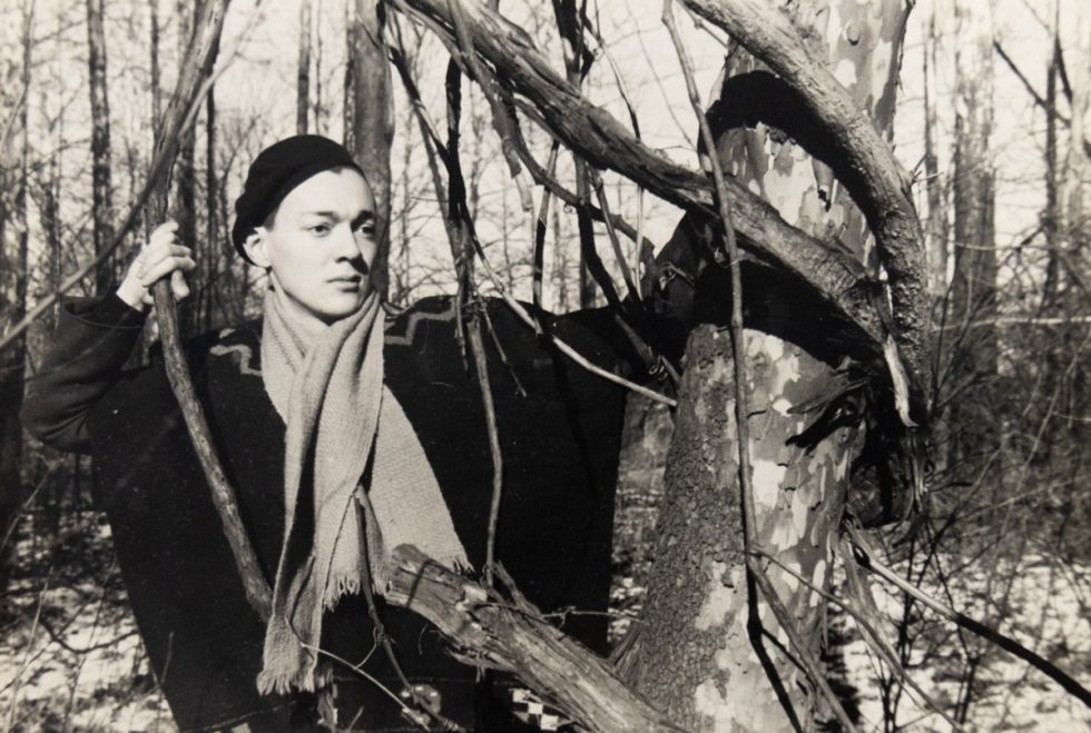 In this black and white image, a person wearing a hat poses in a forest by holding a vine hanging from a tree in each hand.