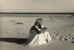 In this black and white photograph, a woman wearing a headscarf sits on a beach with her arms wrapped around her legs. She looks off into the distance.