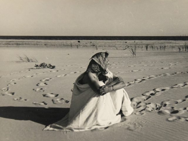 In this black and white photograph, a woman wearing a headscarf sits on a beach with her arms wrapped around her legs. She looks off into the distance.