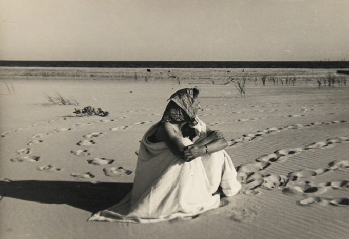 In this black and white photograph, a woman wearing a headscarf sits on a beach with her arms wrapped around her legs. She looks off into the distance.