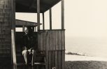 In this black and white image, a woman sits on the porch of a beach house and started off to the left.