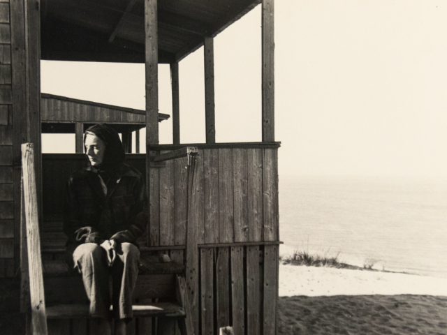 In this black and white image, a woman sits on the porch of a beach house and started off to the left.