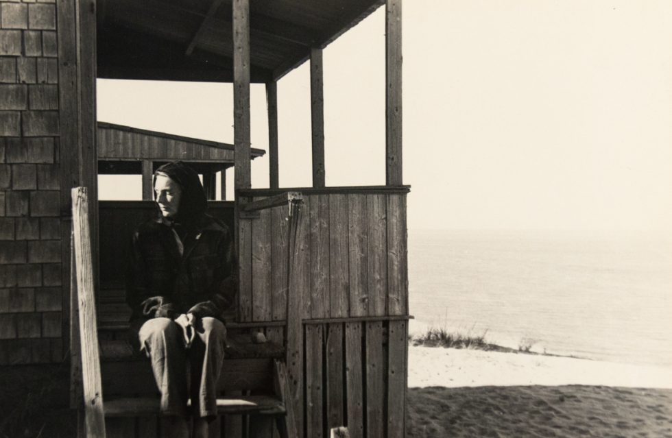 In this black and white image, a woman sits on the porch of a beach house and started off to the left.