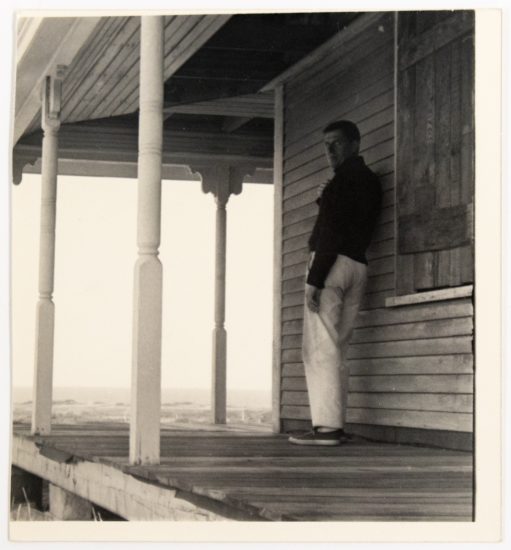 In this black and white image, a man leans against a house while standing on a porch overlooking a bright white sky.