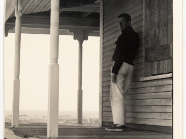 In this black and white image, a man leans against a house while standing on a porch overlooking a bright white sky.