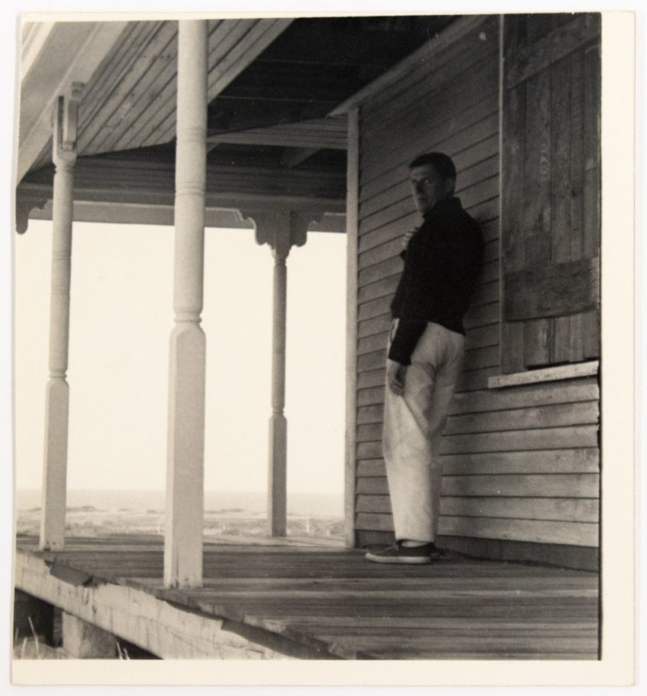 In this black and white image, a man leans against a house while standing on a porch overlooking a bright white sky.