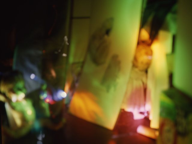 This color photograph shows a blurry space that looks like a kitchen. The figure of a large doll stands next to a white fridge.
