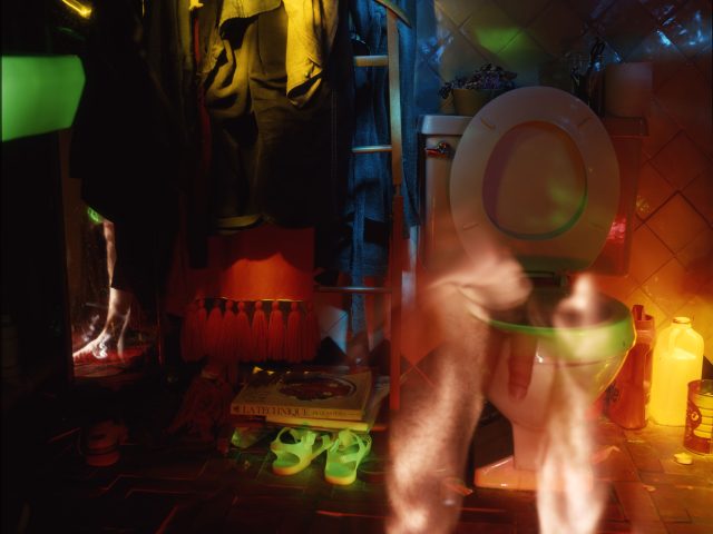 This photograph shows the ghostly figure of a naked man standing in front of the toilet. A pair of green candles lies on the ground off to the left.