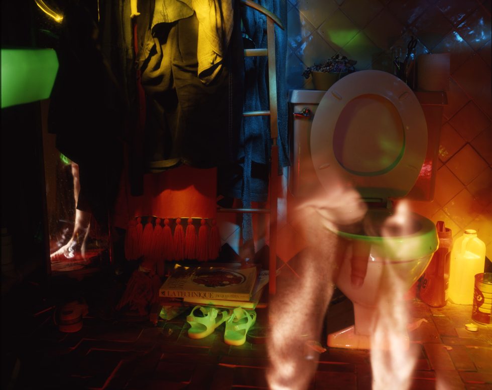 This photograph shows the ghostly figure of a naked man standing in front of the toilet. A pair of green candles lies on the ground off to the left.