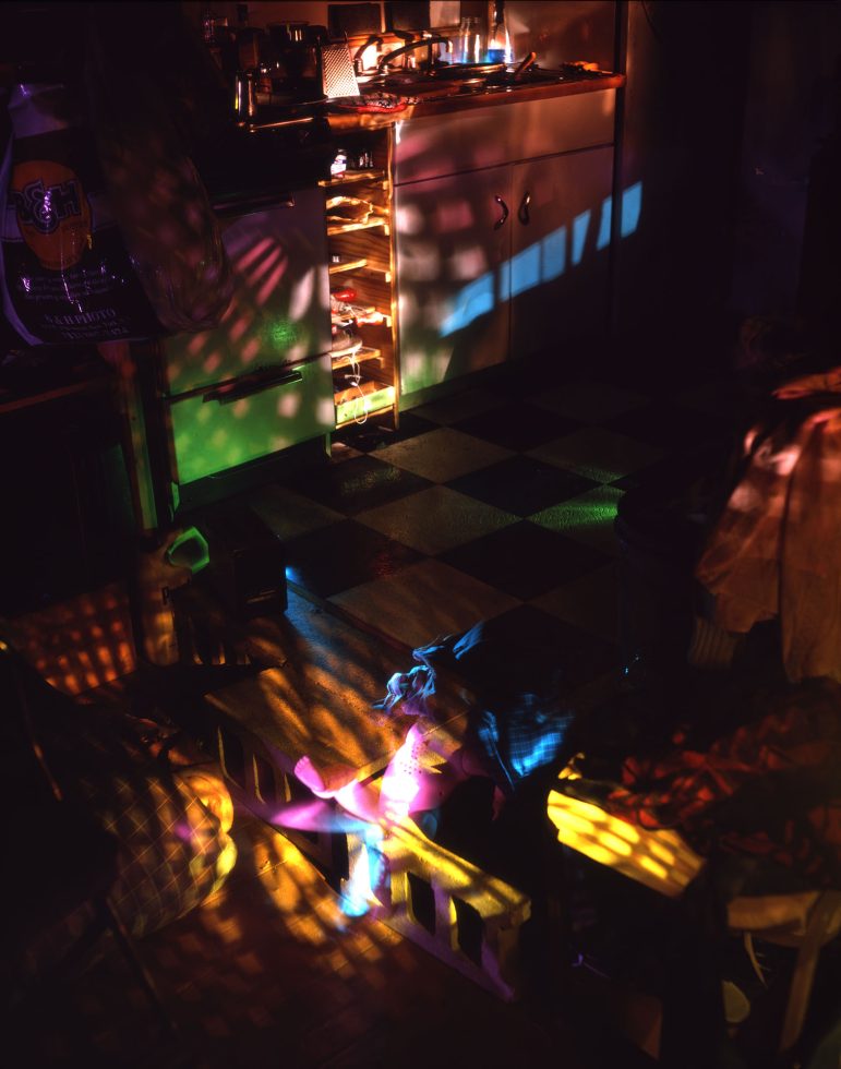 This color photograph shows a kitchen, mostly dark, but lit with scattered rainbows cast across the floor.