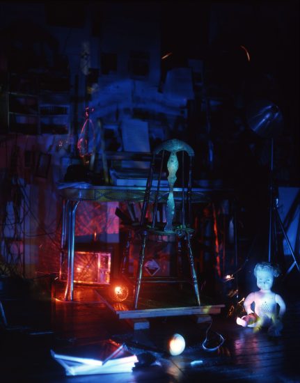 This color photograph shows a bedroom that is glowing with blue light. A doll sits off to the right, below a small desk with a wooden chair.