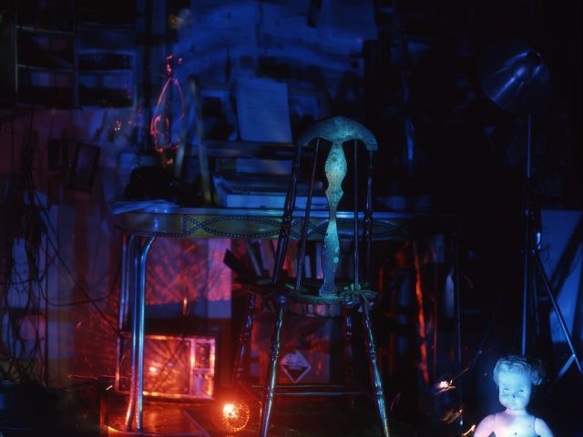 This color photograph shows a bedroom that is glowing with blue light. A doll sits off to the right, below a small desk with a wooden chair.