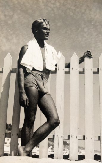 In this vertical black and white photograph, a young man leans against a fence with a white towel wrapped around his shoulders.