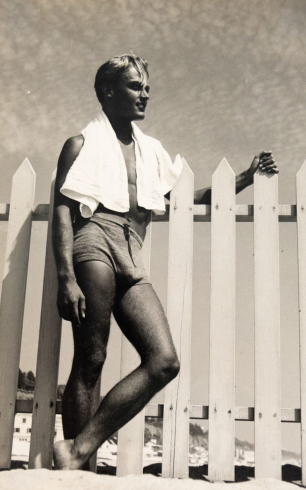 In this vertical black and white photograph, a young man leans against a fence with a white towel wrapped around his shoulders.