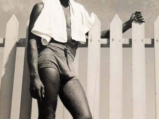 In this vertical black and white photograph, a young man leans against a fence with a white towel wrapped around his shoulders.