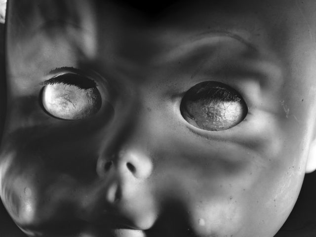 This photograph shows the face of a doll, seen close up. It's eyes are cloudy and set outside its tiny nose.
