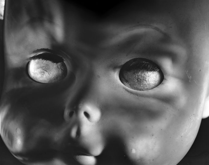 This photograph shows the face of a doll, seen close up. It's eyes are cloudy and set outside its tiny nose.