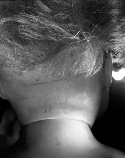 This black and white photograph shows the back of a doll's neck, seen closely. Its skin folders over to create a line across the neck.