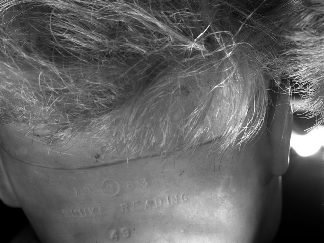 This black and white photograph shows the back of a doll's neck, seen closely. Its skin folders over to create a line across the neck.
