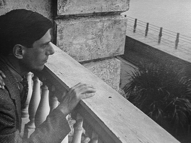 In this black and white photograph, a man looks over the railing of a building. His hand rests on it.