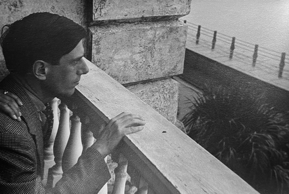 In this black and white photograph, a man looks over the railing of a building. His hand rests on it.