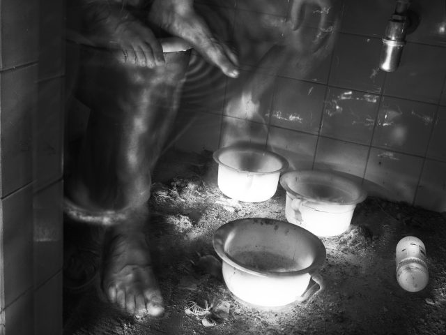 This black and white photograph shows an old bathroom. The floor is covered in a thick layer of dust and dirt. The feet of a ghostly figures sitting behind a wall are floating off to the left.